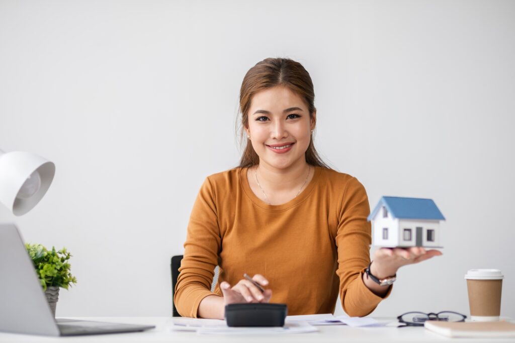 Woman Calculating Real Estate Transaction with Model House and Calculator in Modern Office Setting