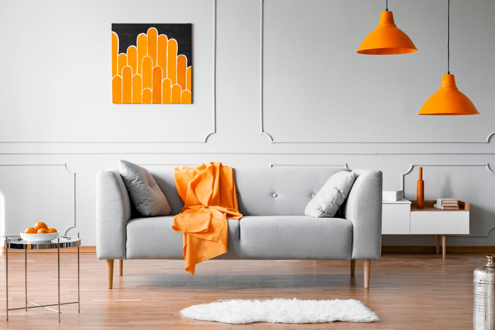 Orange blanket on grey couch in bright flat interior with lamps and poster on the wall. Real photo