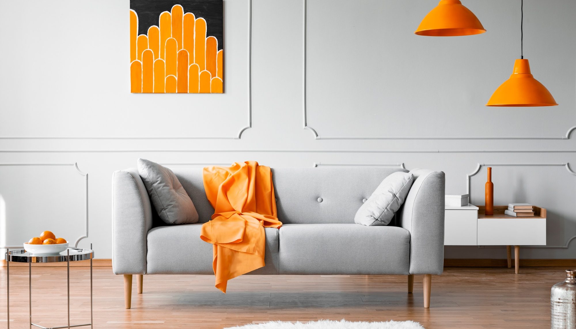 Orange blanket on grey couch in bright flat interior with lamps and poster on the wall. Real photo